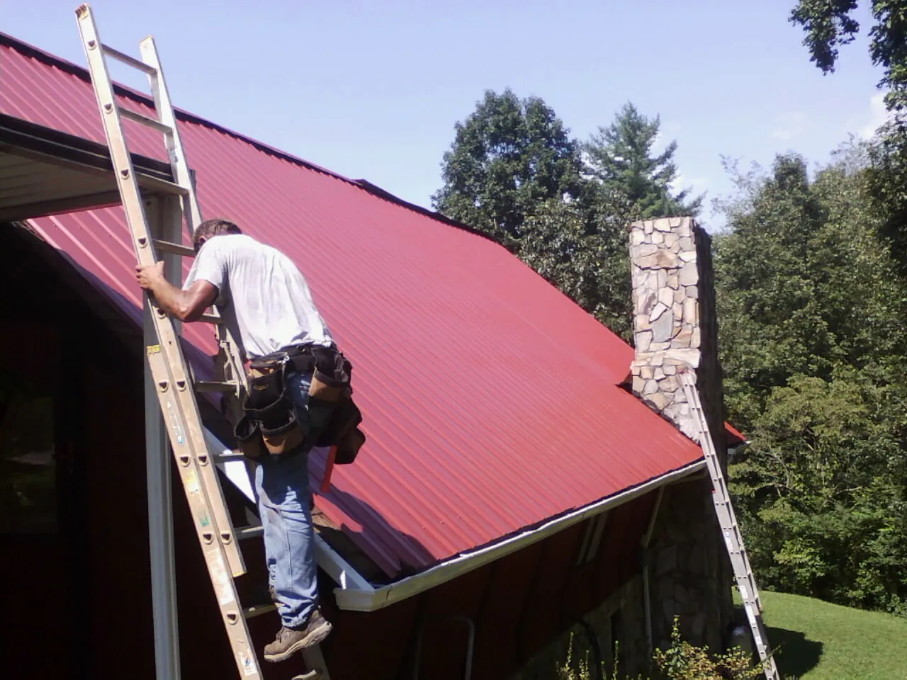 Roofer on a ladder beside a red metal roof and stone chimney — Soffit & Fascia Repair by Roofing Brothers in Homer
